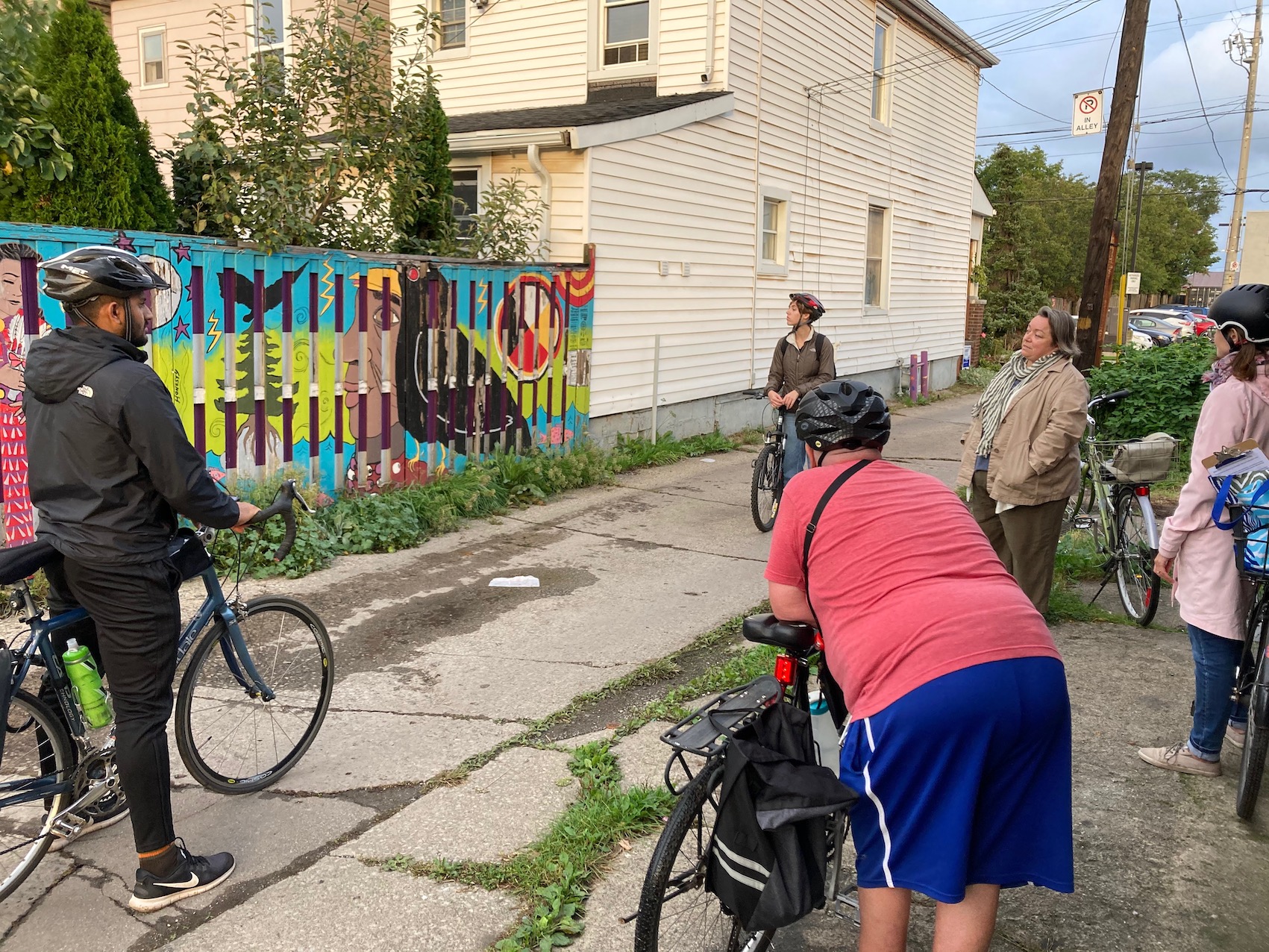 A group of 5 people with bikes listening to someone speak about a colourful mural painted on the background fence