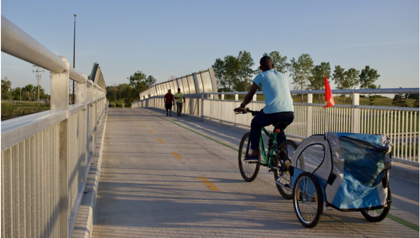 A person cycling over a bridge, captured from behind. They have a trailer attached to their bike.