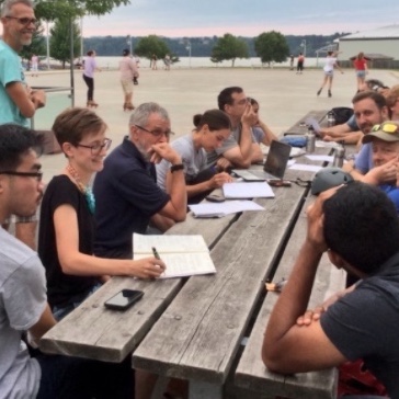 A group of about 10 people sitting around a picnic table outside having a discussion